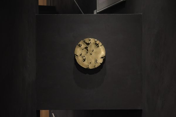Wandrelief von Claudia Barcheri, „Limonade“ (2021): runder, gold-/bronzefarbener Metallkörper mit organischen Durchbrüchen, zentral auf dunkler Wand montiert; gerichtetes Licht wirft Schatten.